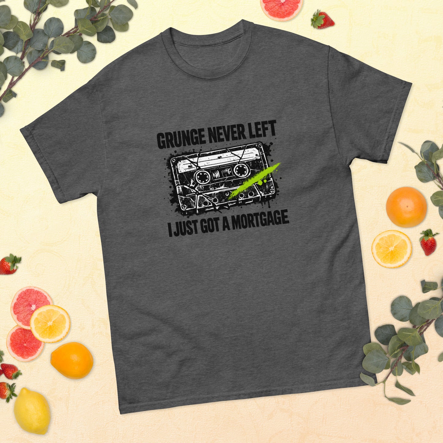 Flat lay mockup of a dark heather gray unisex t-shirt surrounded by sliced citrus fruit, strawberries, and greenery, featuring a 90s cassette graphic with the text "Grunge Never Left I Just Got a Mortgage"
