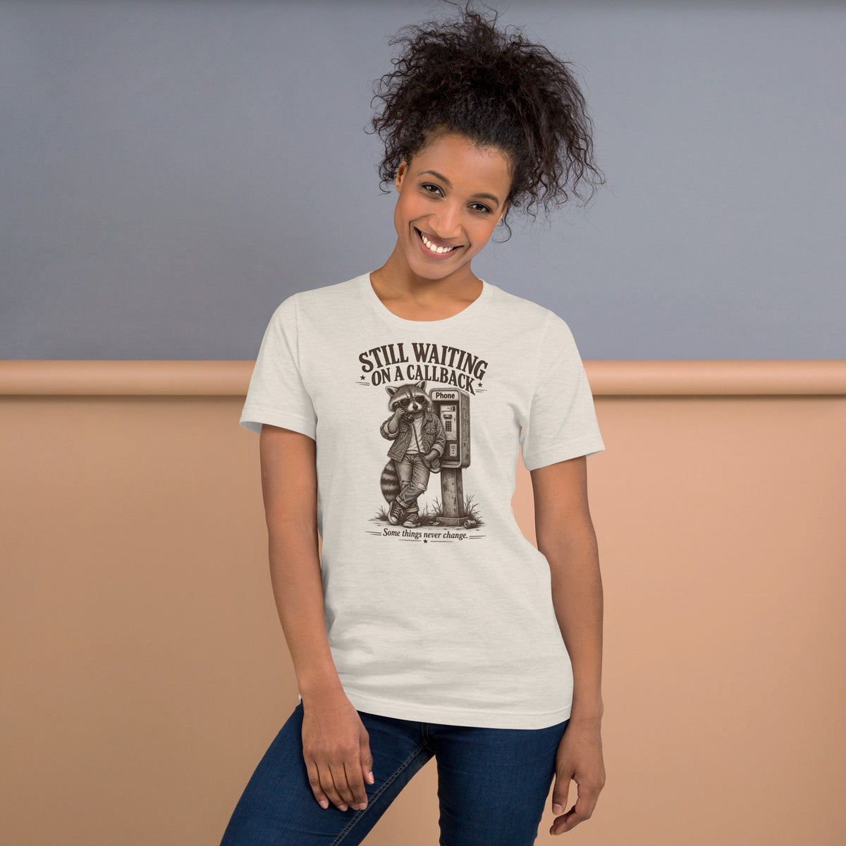 Smiling woman wearing Still Waiting on a Callback funny raccoon graphic t-shirt in beige paired with jeans