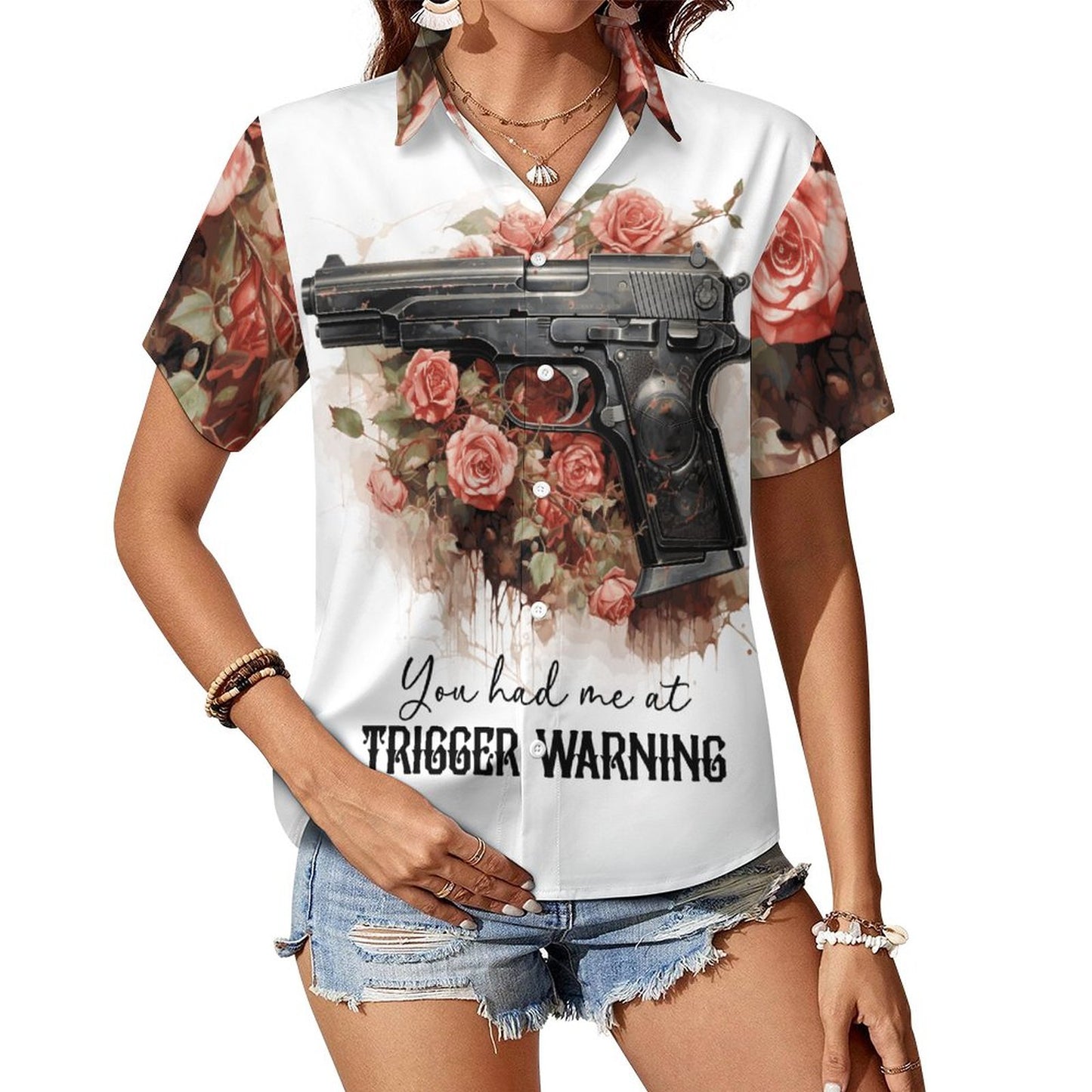 Woman wearing the 'You Had Me at Trigger Warning' short-sleeve button-down shirt paired with denim shorts. The white shirt features an all-over dark romance pattern of pink roses and a handgun graphic design on the front.