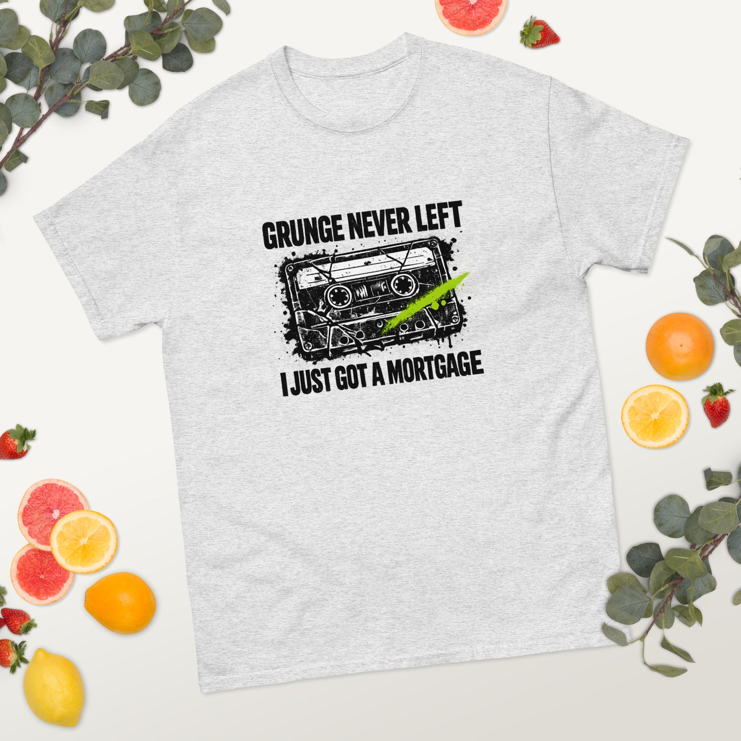 Flat lay mockup of an ash gray unisex t-shirt surrounded by sliced citrus fruit and greenery, featuring a 90s cassette graphic with the text "Grunge Never Left I Just Got a Mortgage"
