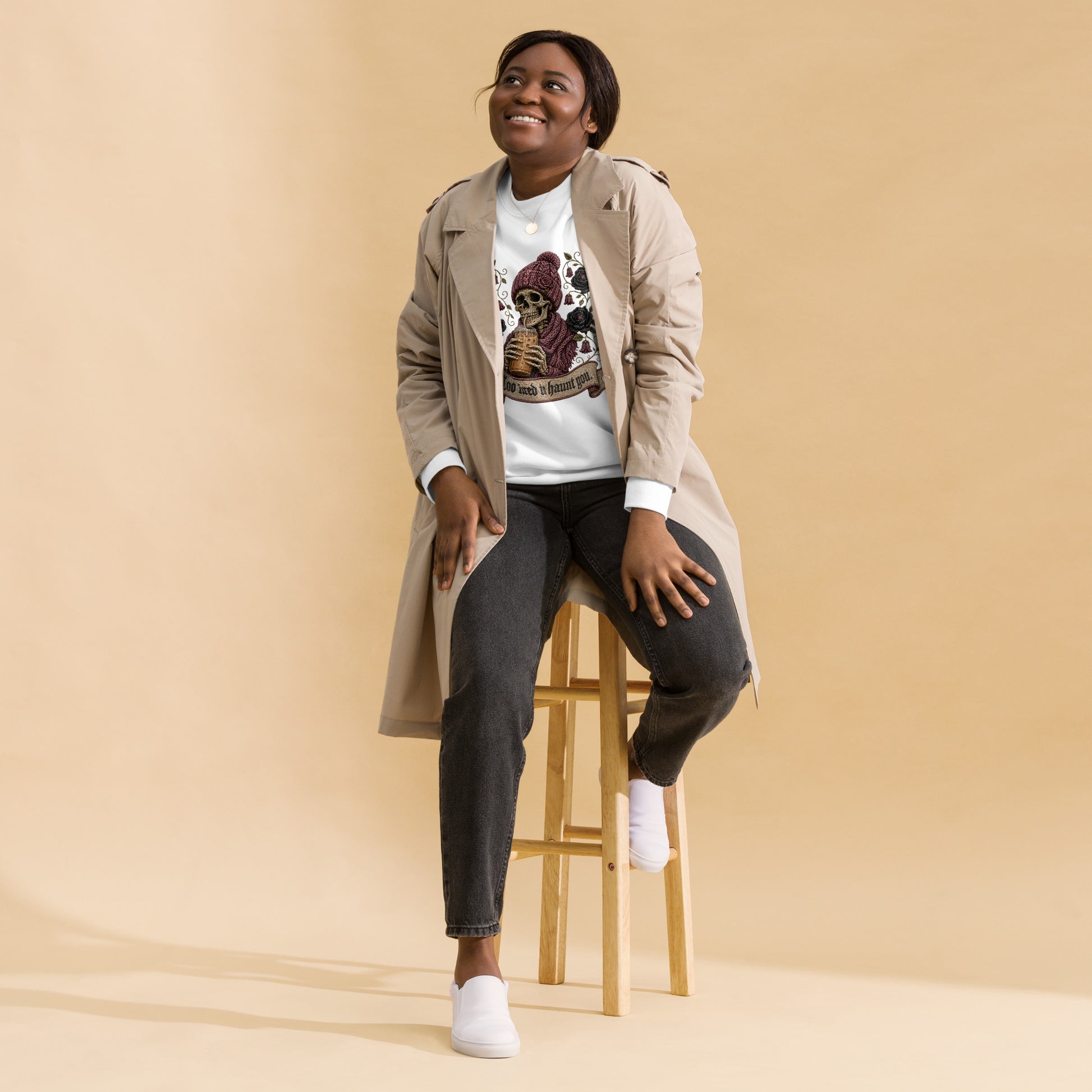 Female model wearing a latte beige unisex crewneck sweatshirt featuring a gothic skeleton in a beanie drinking iced coffee with dark roses and banner reading "Too tired to haunt you"