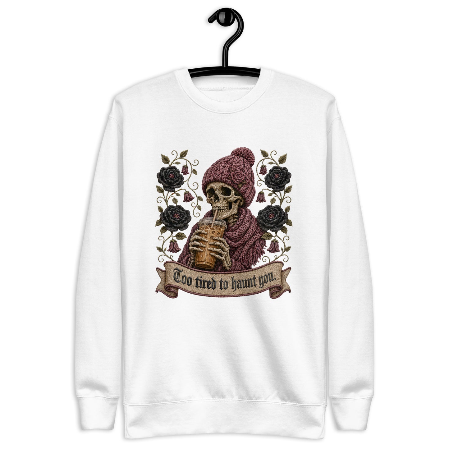 White unisex crewneck sweatshirt hanging on a black hanger featuring a gothic skeleton wearing a beanie drinking iced coffee with dark roses and banner reading "Too tired to haunt you"