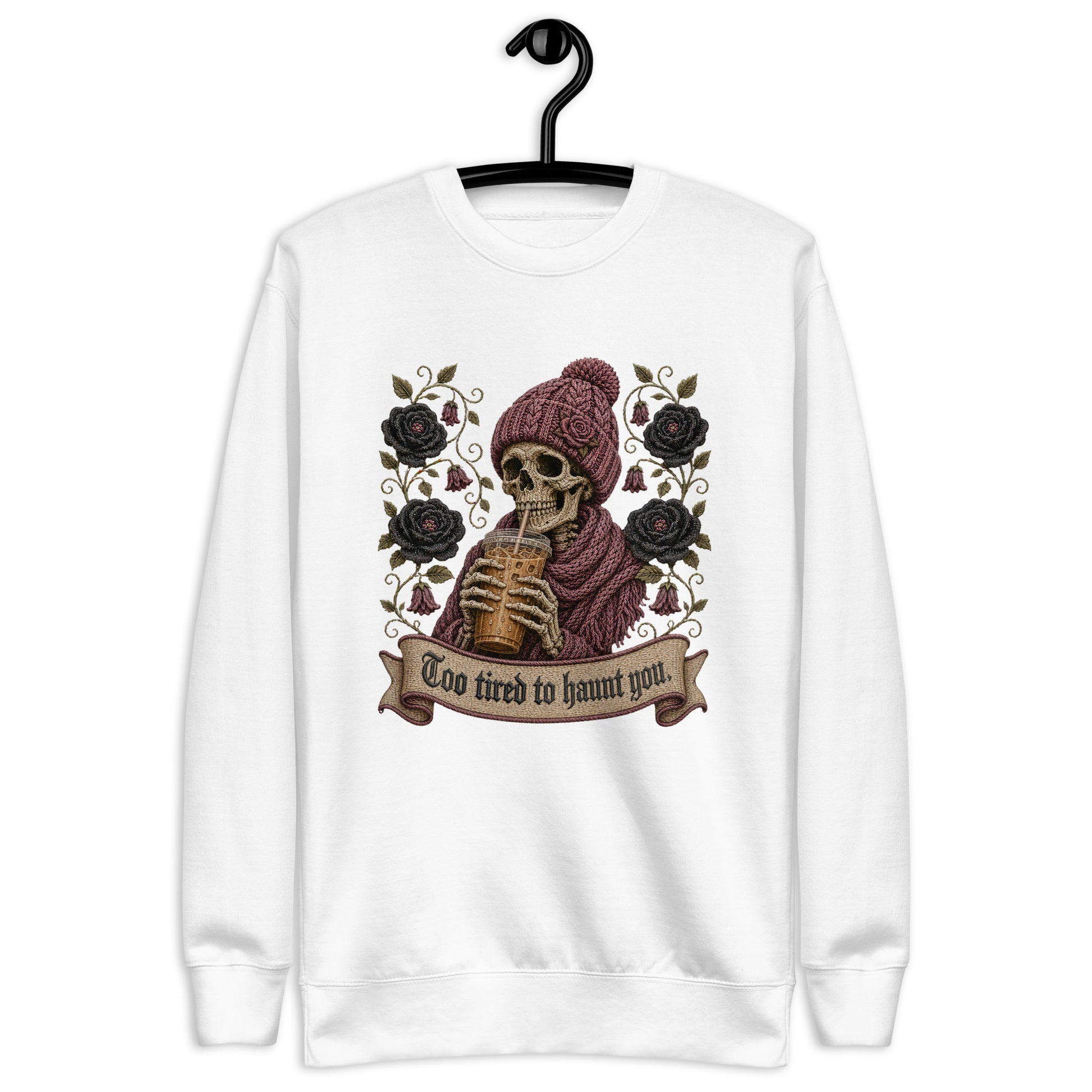 White unisex crewneck sweatshirt hanging on a black hanger featuring a gothic skeleton wearing a beanie drinking iced coffee with dark roses and banner reading "Too tired to haunt you"