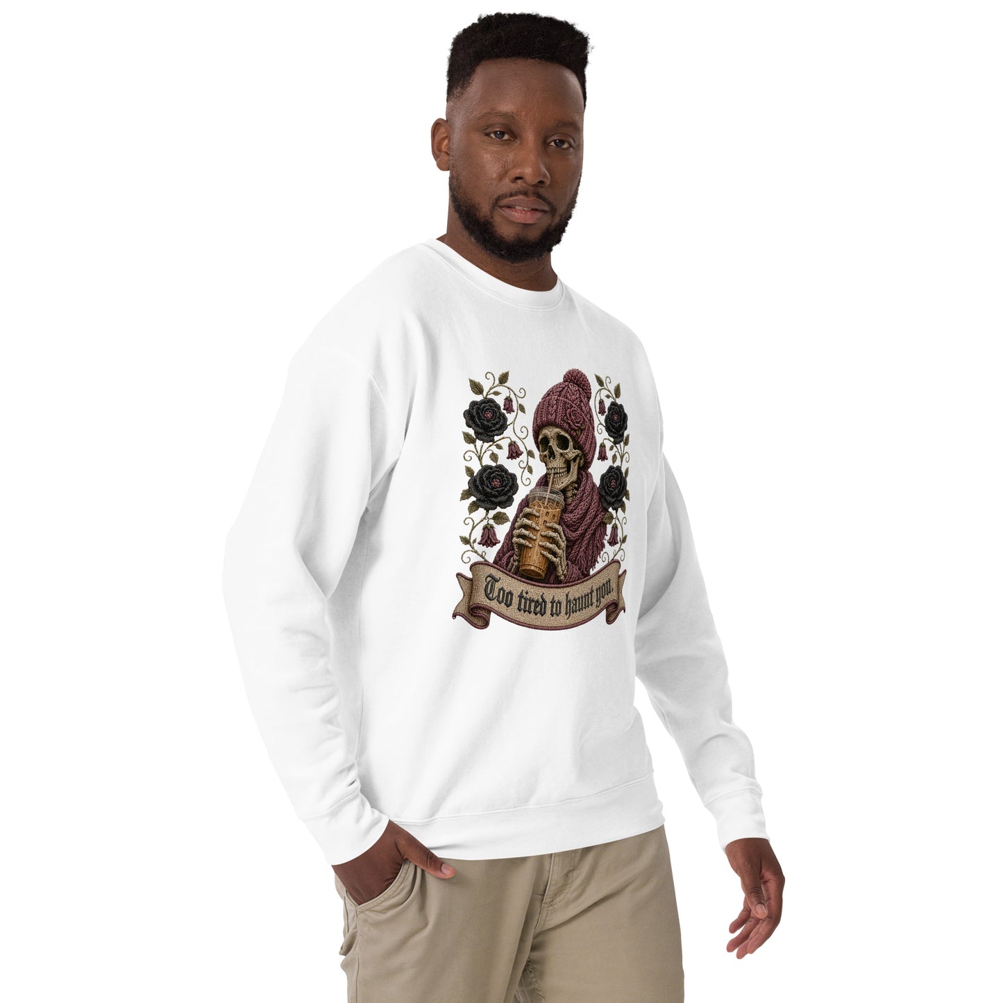 Male model wearing a white unisex crewneck sweatshirt featuring a gothic skeleton in a beanie drinking iced coffee with dark roses and banner reading "Too tired to haunt you"