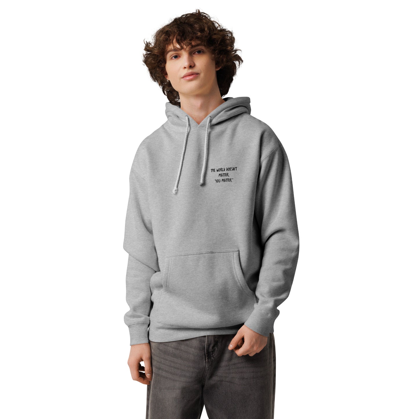 Front view of a model with curly hair wearing the grey heather "in my burgah" unisex heavyweight hooded sweatshirt, featuring small minimalist text graphic on the left chest.